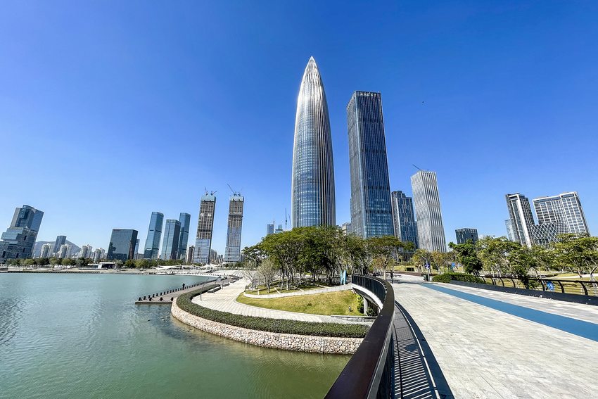 Towers, planting and a waterside walkway at Shenzhen Bay beneath a blue sky.
