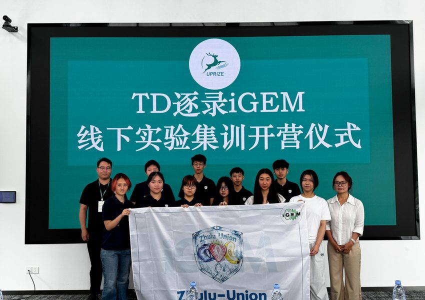Participants pose in front of a screen announcing an experimental training camp, holding a banner with an iGEM mark.