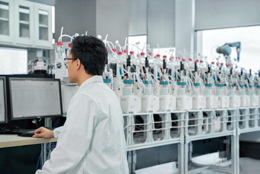 A person in a white laboratory coat works at a computer beside a row of instruments and tubing.