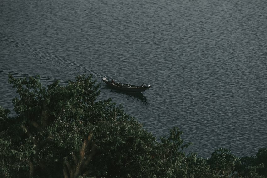 Viewed from above, a narrow boat passes near shoreline trees on calm water.
