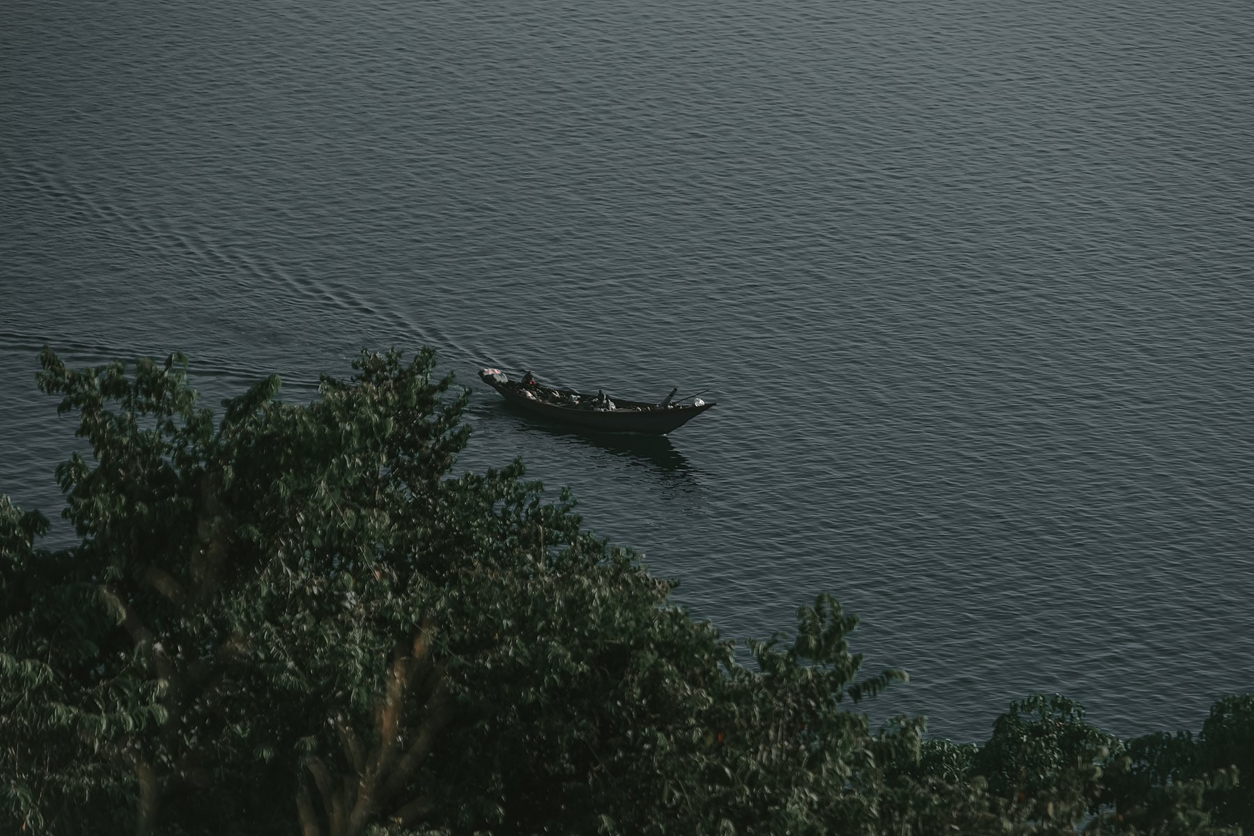 Viewed from above, a narrow boat passes near shoreline trees on calm water.