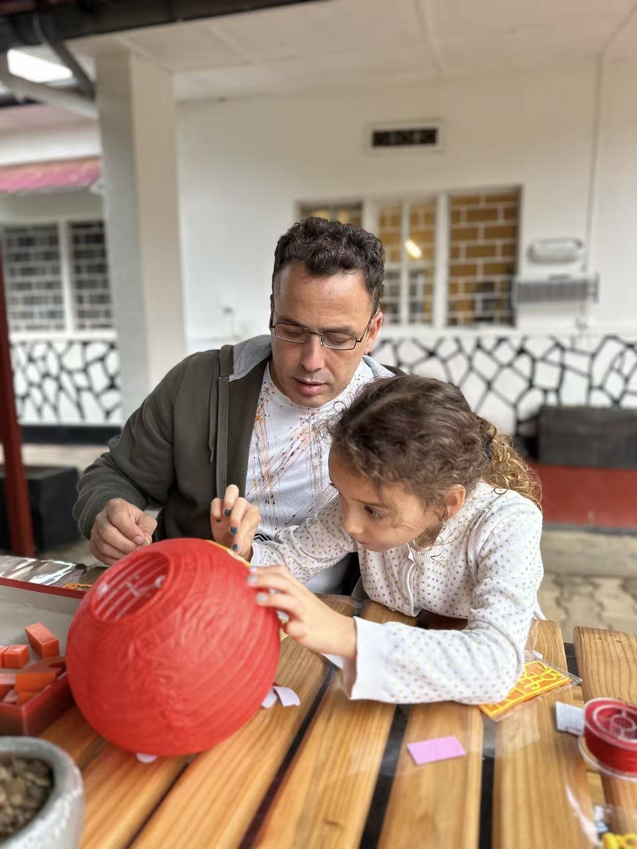 An adult supports a child working on a red lantern at a table.