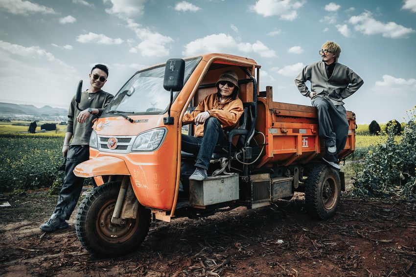 Travellers gather around an orange three-wheeled vehicle beside flower fields.